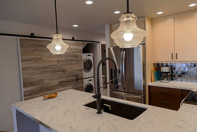 View from kitchen counter with 2 pendant lights hanging from above. Barn door can be seen in the background which opens to a stacked laundry machines.